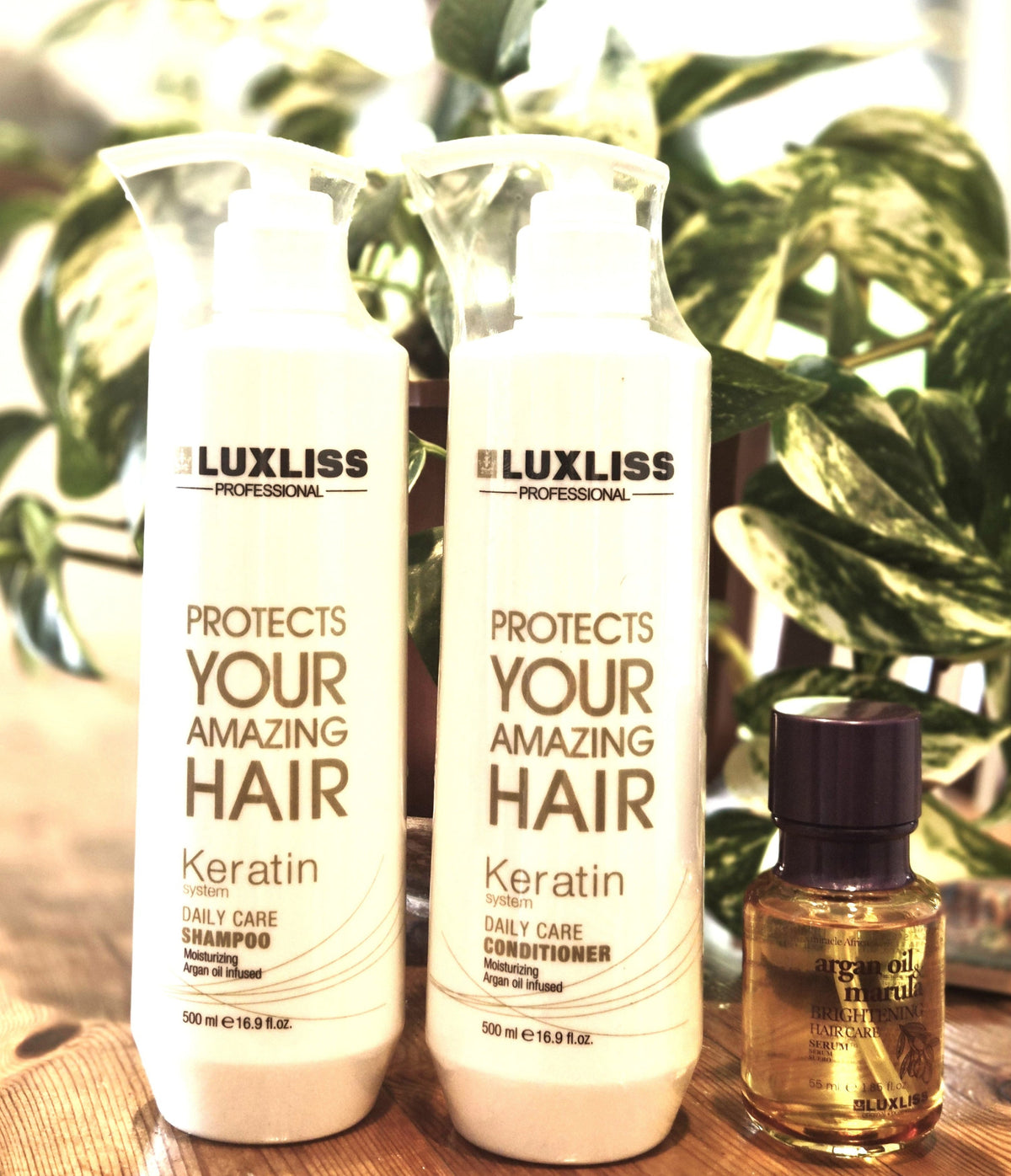 Two white bottles of Luxliss Professional Keratin Daily Care products standing on a wooden surface, with a small amber bottle beside them and green plant leaves in the background.