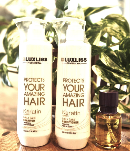 Two white bottles of Luxliss Professional Keratin Daily Care products standing on a wooden surface, with a small amber bottle beside them and green plant leaves in the background.